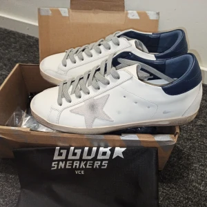 Golden Goose vita sneakers med stjärna - Snygga vita sneakers från Golden Goose med grå mockastjärna på sidan, grå snörning och mörkblå hälkappa. Skorna har platt sula och är tillverkade i skinn med detaljer i mocka. Perfekta för dig som gillar en clean men ändå unik look.