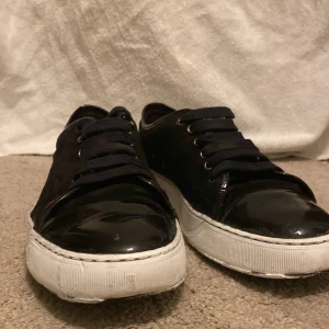 Svarta lanvins  - Snygga svarta sneakers med blanka detaljer och vit, platt sula. Skorna har svarta snören och metallfärgade öljetter. Ovandelen är i syntetmaterial med en glansig finish på tån och hälpartiet. Perfekta för en clean och stilren look. Under sidan är lite dålig men man kan fixa upp den så blir den trevlig igen.