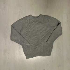 Grå stickad tröja från Maison Margiela - Snygg grå stickad tröja från Maison Margiela med klassisk rund hals och ribbade muddar. Tröjan har ett diskret signaturstygn i nacken och är tillverkad i mjukt material som passar perfekt för kyliga dagar. Enkel och stilren design som funkar till allt.