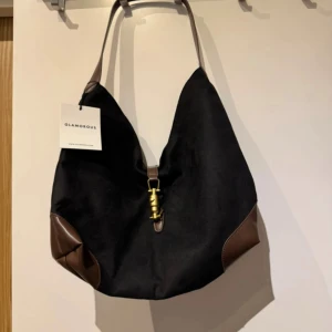 Svart axelväska från Glamorous - Stilren svart axelväska från Glamorous med bruna skinn-detaljer i hörnen och på handtaget. Väskan har en mjuk, slouchy form och ett snyggt guldfärgat spänne framtill. Invändigt finns en praktisk ficka med dragkedja och extra fack för småsaker.
