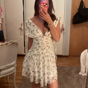 Blommig kortklänning från NA-KD - Supersöt kortklänning från NA-KD i vit med små blommor i rosa, rött och grönt. Klänningen har v-ringning, omlottkänsla upptill, rynkade detaljer i midjan och axelband som knyts. Lätt och flowy i polyester, perfekt för sommarens alla tillfällen. när jag köpte den så fanns det Fläckar på den så har aldrig haft på mig den ute 😞 men jätte fin klänningen 