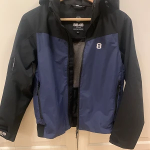 Blå och svart regnjacka 8848 Altitude, stl 150 - Snygg regnjacka från 8848 Altitude i blått och svart med huva och dragkedja framtill. Jackan har två sidofickor med dragkedja, meshfoder och logga på bröstet. Perfekt för dig som gillar stilren och sportig look. Mycket lite använd, fint skick. 