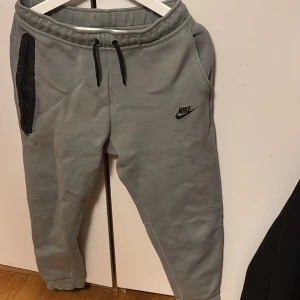 Grå mjukisbyxor från Nike XL för barn - Grå mjukisbyxor från Nike i storlek XL för barn med svart Nike-logga på låret. Byxorna har resår och snörning i midjan, ribbade muddar vid bensluten och en svart dragkedjeficka på ena sidan. Perfekta för chill eller träning.