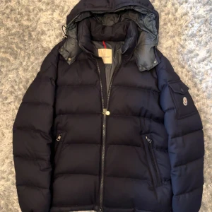 Moncler Montgenevre 2020 pufferjacka med huva - Svart pufferjacka från Moncler med huva, dragkedja och fickor med zip. Jackan har ett broderat Moncler-märke på ärmen och coola detaljer som spännen på sidan. Perfekt för dig som vill ha en stilren och varm jacka med premiumkänsla. Flaws finns, tejpade med nylon för synen samt dunets skull.