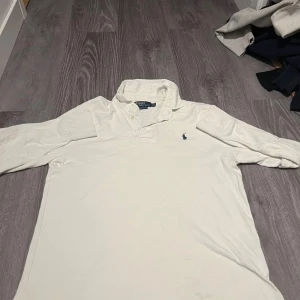 Vit henley från Polo Ralph Lauren - Vit henley från Polo Ralph Lauren med klassisk krage och två knappar vid halsen. Custom fit-modell med broderad marinblå logga på bröstet. Skjortan är långärmad och tillverkad i mjuk bomull, perfekt för en clean och stilren look. 
