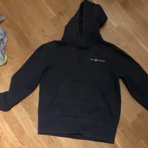 Mörkblå hoodie från Sail Racing - Mörkblå hoodie från Sail Racing med diskret logga på bröstet och en cool detalj på huvan. Tröjan har en stor magficka och mjukt, borstat foder på insidan. Perfekt för dig som gillar enkel och stilren streetwear. Köpt från kidsbrandstore och knappt använd.