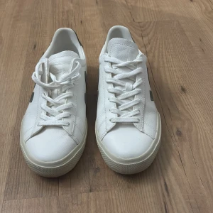 Vita Veja sneakers i skinn - Snygga vita sneakers från Veja med klassisk låg profil. Skorna är i skinn med ”khaki” färgade mockadetaljer på hälen och den ikoniska V-loggan på sidan. De har rund tå, platt sula och snörning framtill. Perfekta för en clean och stilren look.