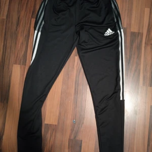 Svarta Adidas träningsbyxor S - Svarta träningsbyxor från Adidas i storlek S med klassiska vita ränder längs sidorna och Adidas-logga på låret. Byxorna har elastisk midja och är tillverkade i ett mjukt, syntetiskt material som passar perfekt för sport och träning.