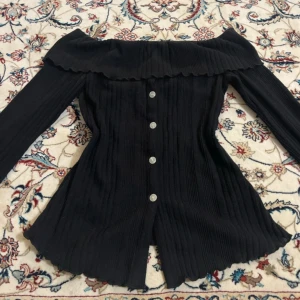 Svart offshoulder topp - Säljer en svart offshoulder topp med långa ärmar och ribbad struktur. Toppen har dekorativa knappar framtill och en vågig kant längs halsringningen och nederdelen. Materialet är mjukt och stretchigt, perfekt för att ge en snygg siluett.