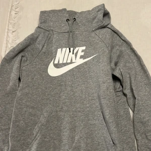 Grå hoodie från Nike med logga - Säljer en grå hoodie från Nike med stor vit logga på bröstet. Tröjan har huva med snörning och en klassisk känguruficka framtill. Mjuk och skön bomullsblandning, perfekt för chill eller träning. Långärmad och normal passform.