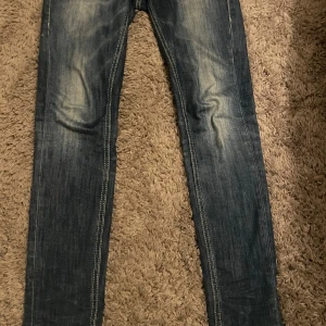 Blå skinny jeans från Dondup - Säljer ett par blå skinny jeans från Dondup med snyggt slitna detaljer och kontrastsömmar. Jeansen har klassisk femficksdesign och är tillverkade i mjukt denimtyg som sitter tajt längs benen. Perfekta för dig som gillar en smal siluett.