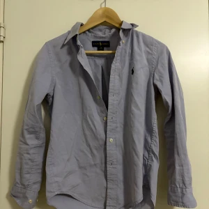 Ljusblå skjorta från Ralph Lauren - Klassisk ljusblå skjorta från Ralph Lauren med lång ärm och knappar framtill. Skjortan har en broderad logga på bröstet och är i en custom fit-modell. Perfekt för en stilren och avslappnad look.
