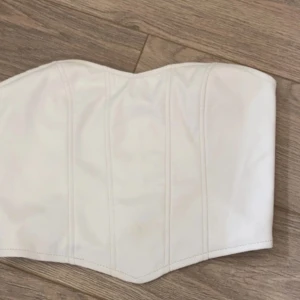 Vit korsett-topp i syntetmaterial - Säljer en vit korsett-topp med snygg formad kant upptill och markerade sömmar framtill. Toppen är axelbandslös och har en stilren look som är lätt att matcha. Materialet är slätt och känns syntetiskt, perfekt för dig som gillar trendiga och cleana plagg.
