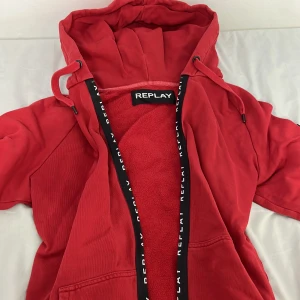 Röd hoodie från Replay med huva - Röd hoodie från Replay med dragkedja och svart logoband längs öppningen. Tröjan har huva med snörning, mjuk fleece på insidan och en patch med Replay-logga på ärmen. Perfekt för chill dagar och streetstyle.