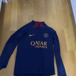 PSG långärmad träningströja Nike M - Paris Saint-Germain långärmad träningströja från Nike i marinblått med vinröda detaljer vid kragen. Slim fit-modell med guldfärgade tryck, inklusive Qatar Airways och PSG-logga på bröstet. Tillverkad i lätt och funktionellt material, perfekt för fotbollsträning.