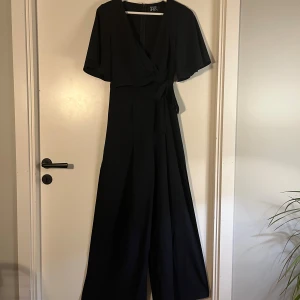 Svart byxdress från Parallel Lines XS - Snygg svart byxdress/overall från Parallel Lines i storlek XS. Modellen har korta, vida ärmar, omlott framtill med knyt i midjan och vida ben, öppna på sidan. Materialet är mjukt och faller snyggt, dragkedja bak för enkel påklädning. Perfekt för ett speciellt tillfälle typ en gala.