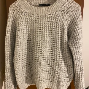 Grå stickad tröja från 157 - Mysig grå stickad tröja från 157 med grovstickat mönster och rund halsringning. Tröjan har långa ärmar och ribbade muddar vid ärmslut och nederkant. Perfekt för kyliga dagar och enkel att matcha med jeans eller kjol.