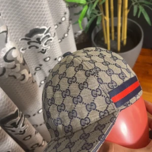 Gucci keps med GG-mönster och röd-blå rand - Snygg keps från Gucci i beige med klassiskt GG-monogram i mörkblått över hela kepsen. På sidan finns en ikonisk röd och marinblå rand. Kepsen har justerbar rem baktill och är fodrad med mörkblått tyg. Perfekt för dig som vill sticka ut med lyxig streetstyle.