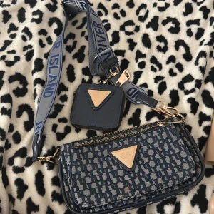 Blå axelväska från River Island - Snygg blå axelväska från River Island med matchande liten plånbok. Väskan har ett mönstrat tyg med River Island-logga, guldfärgade detaljer och dragkedjor. Justerbar axelrem med logotyp och flera fack inuti för smart organisering.