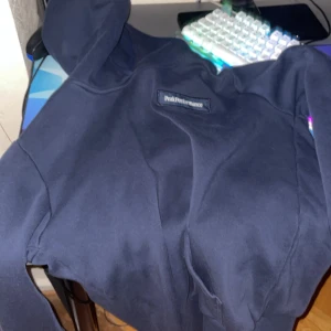 Mörkblå hoodie Peak Performance - Snygg mörkblå hoodie från Peak Performance med klassisk logga på bröstet. Tröjan har huva och är tillverkad i mjukt bomullsmaterial, perfekt för chill dagar. Passar dig som gillar stilrena och sportiga plagg.