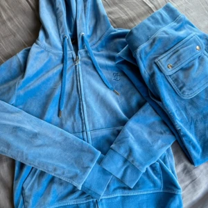 Blå velourbyxor Juicy Couture XS - Säljer ett par ikoniska blå velourbyxor och en tröja från Juicy Couture i storlek XS. Byxorna har breda ben, snörning i midjan och stora fickor framtill med broderad JC-logga. Perfekta för en chill och trendig look.