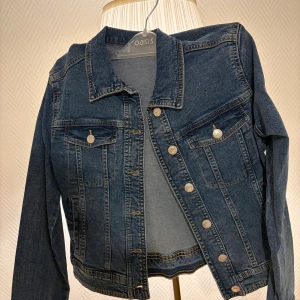 Mörkblå jeansjacka från Oasis - Snygg mörkblå jeansjacka från Zalando med klassisk krage, silverfärgade knappar och två bröstfickor med lock. Jackan har lång ärm och markerade sömmar som ger en cool look. Perfekt att slänga över en hoodie eller t-shirt.
