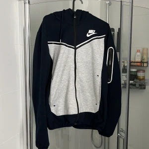 Nike sportig vindjacka med huva - Nike vindtröja i svart och grått med vit logga på bröstet. Tröjan har huva med snörning, hel dragkedja och en cool ficka med dragkedja på ärmen. Sportig design med kontrastpaneler och lång ärm. Perfekt för en aktiv stil.