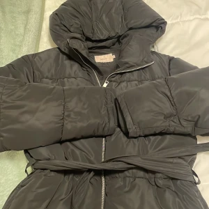 Svart pufferjacka från Nelly XS - Svart pufferjacka från Nelly i storlek XS. Jackan har huva, långa ärmar, dragkedja framtill och ett bälte i midjan för extra stilpoäng. Fickor på sidorna men ena sidan är trasig det går att sy. Perfekt för vinter används 1 gång. Pris kan diskuteras🥰