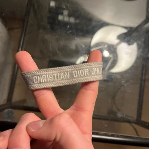 Grått armband Christian Dior - Snyggt grått armband från Christian Dior med broderad logga. Armbandet är gjort i textil och har en stilren, minimalistisk design som passar perfekt till din outfit. Enkel att bära och ger en exklusiv känsla.