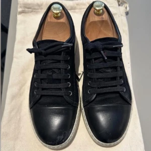 Svarta sneakers från Lanvin - Snygga svarta sneakers från Lanvin med vit platt sula och rund tå. Ovandelen är i mocka med detaljer i skinn på tån och snörning framtill. Klassisk och stilren design som passar perfekt till jeans eller chinos. Märket syns på sulan och medföljer dustbag.