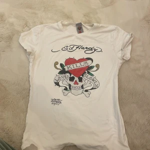 Vit Ed Hardy t-shirt med tryck XL - Vit t-shirt från Ed Hardy i storlek Xl med passar xs med ikoniskt tattoo-inspirerat tryck framtill. Motivet har en dödskalle, hjärta och banderoll med texten 'Love Kills Slowly'. Klassisk passform och korta ärmar. Tillverkad i mjuk bomull. 