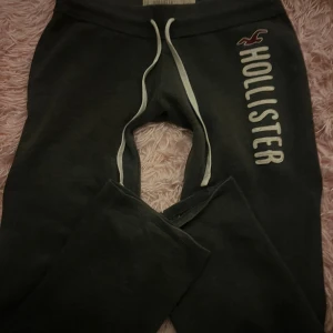 Mörkgrå mjukisbyxor från Hollister - Svarta mjukisbyxor från Hollister med vit snörning i midjan och stort vitt tryck längs benet. Byxorna har en relaxed passform och är gjorda i mjukt bomullsmaterial, perfekta för chill dagar. Klassisk Hollister-logga upptill.