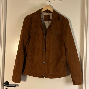 Brun jacka från Cubus, storlek S - Snygg brun sjacka i typ konstmocka från Cubus i storlek S. Jackan har klassisk krage, knappar framtill och två bröstfickor med lock. Materialet är 100% polyester på utsidan och insidan är delvis bomull. Perfekt för dig som gillar stilrena och tidlösa plagg.