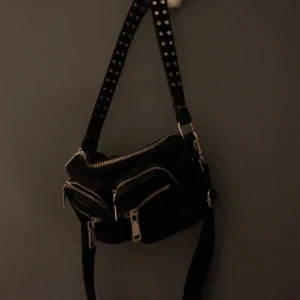 Noella väska svart - Cool svart axelväska från Noella med två framfickor och silvriga dragkedjor. Väskan har bred axelrem med nitar och är tillverkad i mjukt tygmaterial. Perfekt storlek för mobilen, plånboken och andra småsaker.