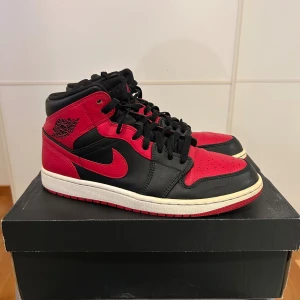 Nike Air Jordan 1 Mid Banned - Säljer ett par Nike Air Jordan 1 Mid Banned i svart och rött skinn med vita detaljer. Använda men fortfarande i hyfsat bra skick. Klassisk siluett med snörning, perforerad tå och ikoniska Jordan Wings-loggan på sidan. Röd yttersula och svart/röd ovandel ger en riktigt clean och sportig look.