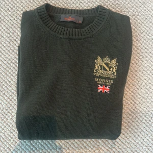 Stickad tröja Morris  - Snygg mörkgrön stickad tröja från Morris med broderat emblem och brittisk flagga på bröstet. Rund halsringning och ribbade muddar vid ärmslut och nederkant. Den är i 100% bomull. Tröjan är nvänd mycket men ändå i fint skick. 