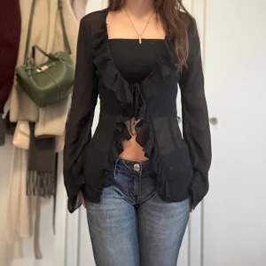Svart volangblus med knytning - Säljer en svart transparent blus med långa ärmar och volangdetaljer längs hela framsidan. Blusen har knytning framtill och är lätt croppad. Perfekt för dig som gillar en trendig och feminin look.