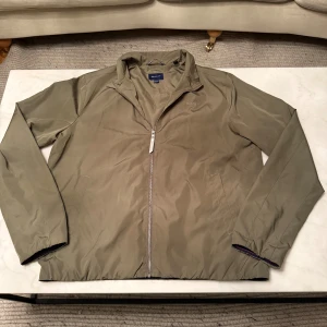 Olivgrön vindjacka från GANT - Snygg olivgrön vindjacka från GANT med dragkedja framtill och diskret logga på bröstet. Jackan har två fickor, ståkrage och elastiska muddar. Perfekt för dig som gillar stilren och sportig look.