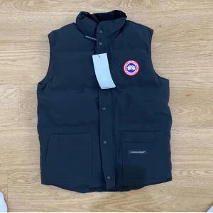 Svart dunväst från Canada Goose - Svart dunväst från Canada Goose med hög krage, två stora fickor framtill och klassisk logotyp på bröstet. Västen har knäppning med tryckknappar och en stilren, enkel design som passar perfekt till lager-på-lager-stil.