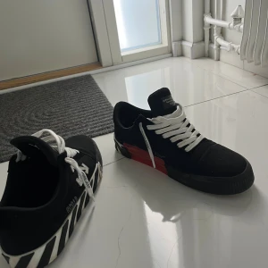 Off-White svarta sneakers med ränder - Säljer ett par svarta Off-White sneakers med vita snören och ikoniska Off-White-detaljer. Skorna har svart och vit randig sula, röd detalj på sidan och klassisk Off-White-logga. Tillverkade i canvas med platt sula och rund tå. Perfekta för dig som gillar streetwear och unika detaljer.