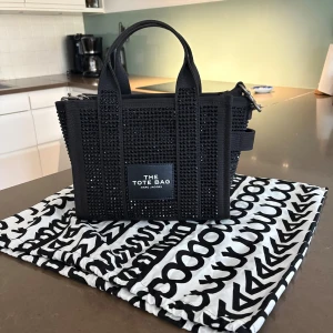 Svart Tote Bag Marc Jacobs med kristaller - Snygg svart Tote Bag från Marc Jacobs dekorerad med svarta kristaller för extra bling. Väskan har både handtag och axelrem, dragkedja upptill och tydlig logga framtill. Perfekt för dig som vill sticka ut med en trendig och cool accessoar. Aldrig använt, prislappen är på.