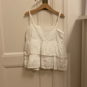 Vit volangtop från Gina Tricot - Säljer en söt vit linnetopp från Gina Tricot med tunna axelband och broderade blommor. Toppen har två lager volanger som ger en luftig och chill vibe. Perfekt för varma dagar och enkel att matcha med jeans eller shorts. Säljer då den inte kommer till användning längre 