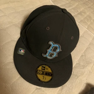 Svart New Era 59FIFTY Boston keps - Snygg svart New Era 59FIFTY keps med Boston B-logga i blått och silver framtill. Officiell MLB On-Field Cap, broderade detaljer och klassisk flat skärm. Tillverkad i 100% polyester för en skön och hållbar känsla.