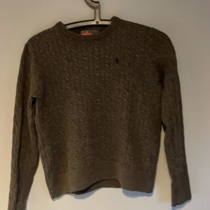 Mörkgrå kabelstickad tröja Morris - Snygg mörkgrå kabelstickad tröja från Morris i 100% merinoull. Tröjan har rund hals, ribbade muddar och ett diskret broderat emblem på bröstet. Perfekt för dig som gillar klassisk stil med en modern touch.