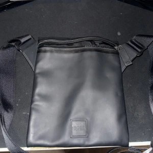 Svart axelväska från Hugo Boss - Stilren svart axelväska från Hugo Boss i slät syntet med justerbar rem och dragkedja upptill. Väskan har minimalistisk design med diskret logotyp framtill och ett rymligt huvudfack. Perfekt för att bära det viktigaste på stan.