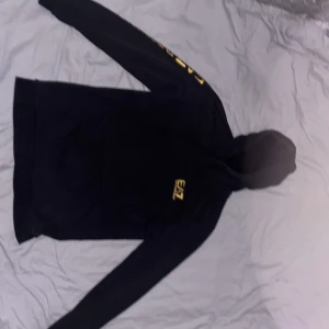 Svart EA7 hoodie med guldtryck - Svart hoodie från EA7 Emporio Armani med guldig logga på bröstet och längs ena ärmen. Tröjan har huva med snörning och en klassisk känguruficka framtill. Perfekt för dig som gillar streetwear och vill sticka ut med exklusiva detaljer. Det finns lite hål vid fickan men inte några stora så därför sätter jag den på lägre pris. Det står i tröjan att den är XXL men den sitter som en S eller M tröja