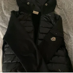 Svart pufferjacka från Moncler - Svart pufferjacka från Moncler med huva och dragkedja. Jackan har quiltade partier och ett snyggt Moncler-märke på ärmen. Materialet är glansig polyester och jackan har en modern, sportig look som passar perfekt till kyliga dagar.