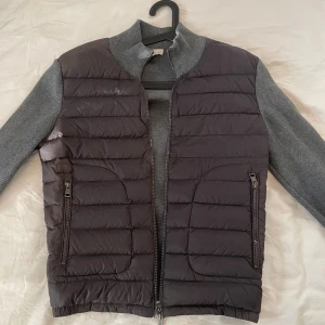 Grå Moncler pufferjacka med stickade ärmar - Snygg pufferjacka från Moncler i mörkgrå färg med stickade ärmar och krage. Jackan har två dragkedjeförsedda fickor och quiltad front. Moncler-logga på ärmen och DIST-märkning på insidan. Perfekt för dig som vill ha en stilren och modern look.