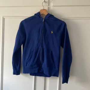 Blå hoodie från Polo Ralph Lauren - Blå hoodie från Polo Ralph Lauren med dragkedja framtill och klassisk gul logga på bröstet. Tröjan har huva, kängurufickor och är tillverkad i mjuk bomull. Perfekt för en chill och avslappnad stil.