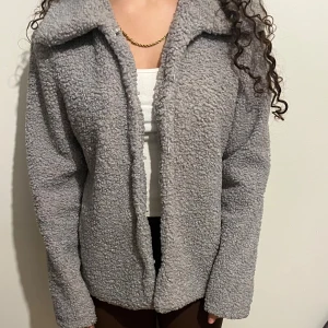 Grå teddyjacka med krage - Mysig grå teddyjacka med bred krage och långa ärmar. Jackan har en loose passform och stängs med diskreta knappar framtill. Materialet är fluffigt och ger en chill vibe. Enkel design utan mönster eller extra detaljer. Väldigt fin och mysig och perfekt att ha på sig nu. 